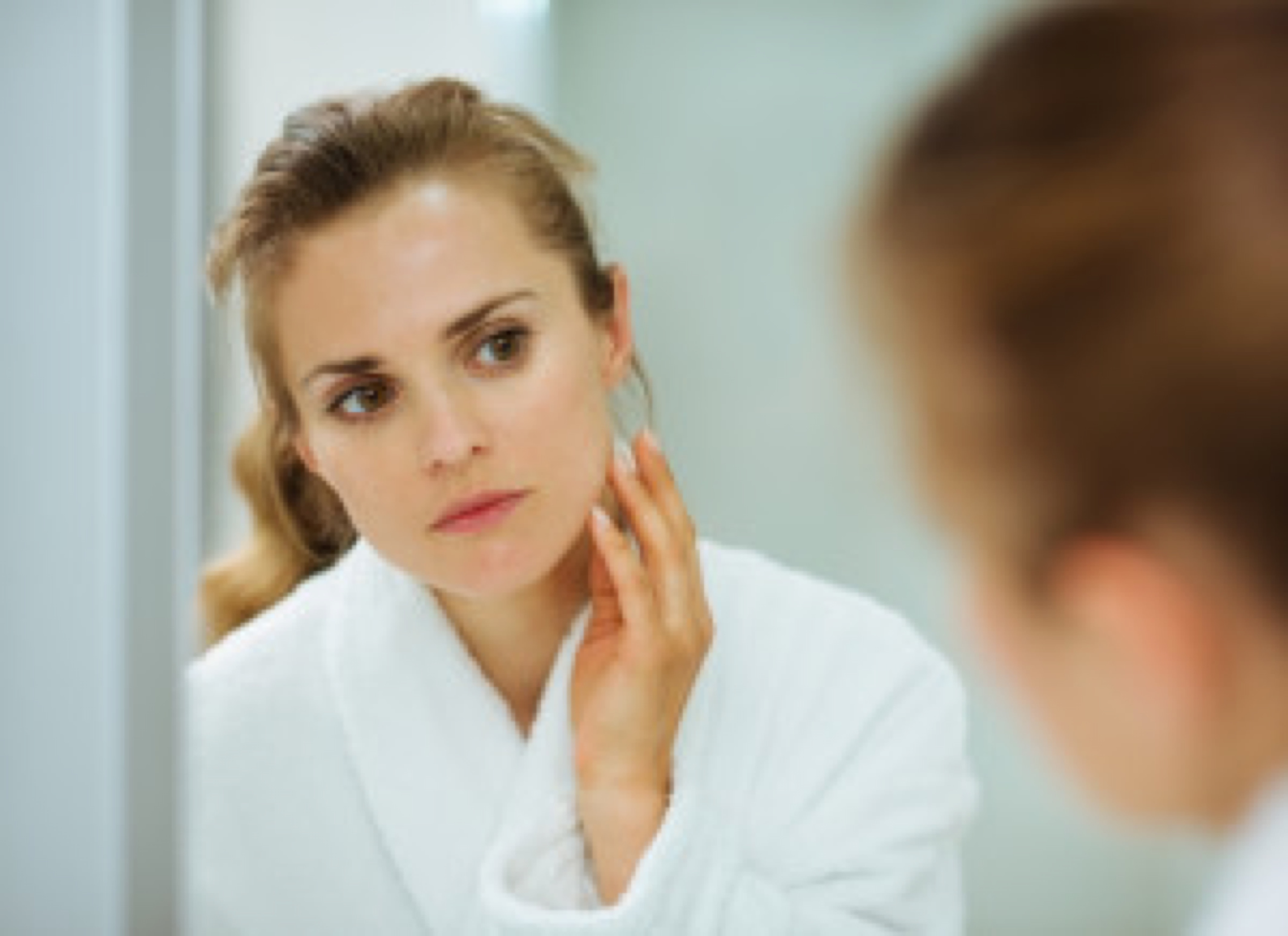 Young woman in bathrobe checking her face in mirror in bathroom