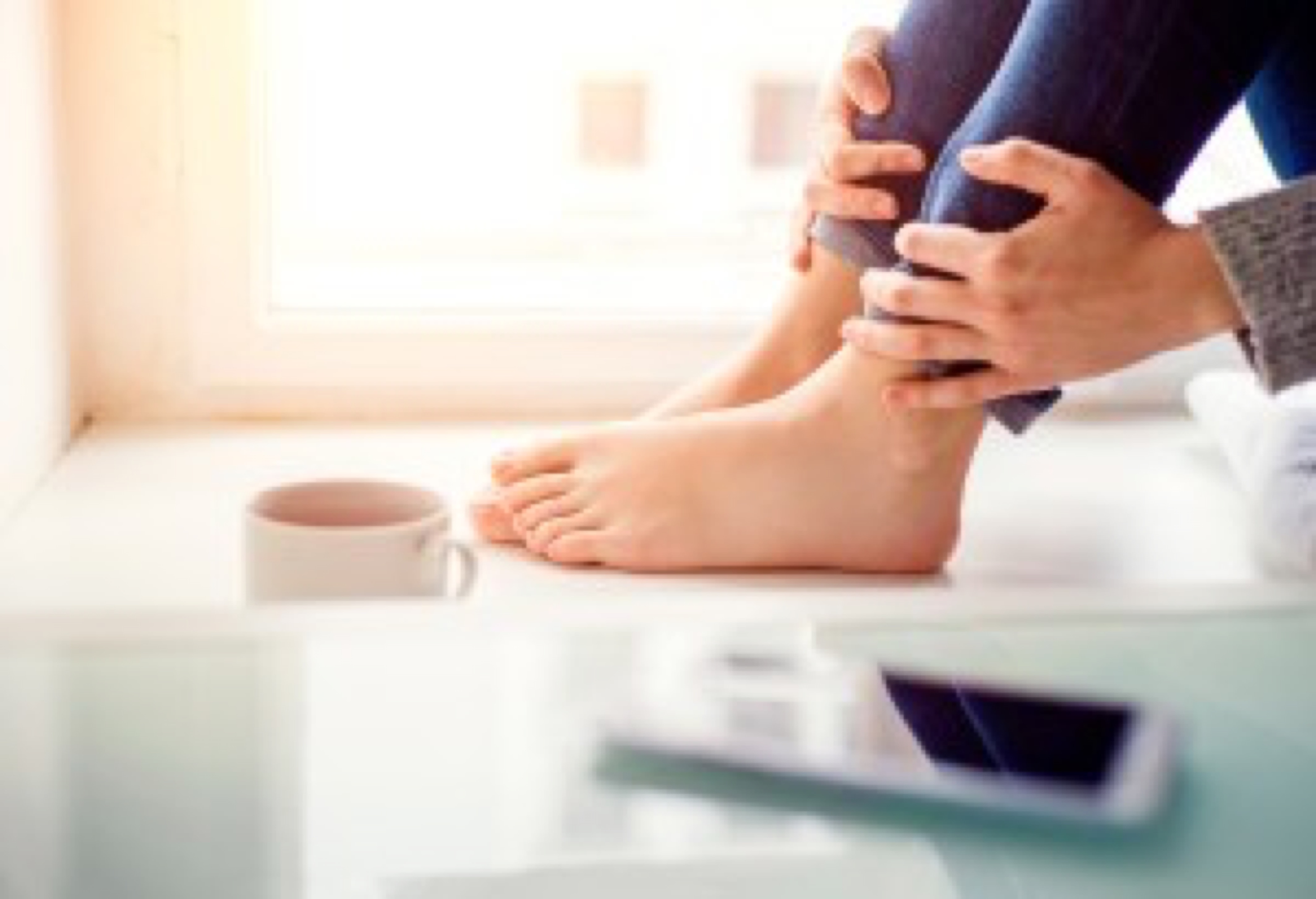 Feet of unrecognizable woman sitting on window sill with smart phone and cup of coffee